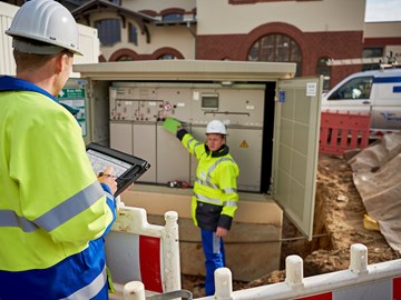 Zwei Techniker in Warnschutzjacken mit portablem Eingabegerät vor offener Trafostation Zwei Techniker in Warnschutzjacken mit portablem Eingabegerät vor offener Trafostation