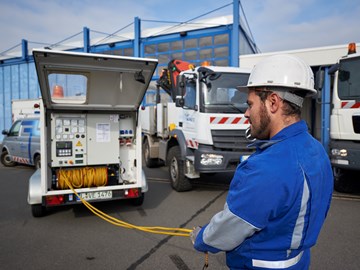 Mitarbeiter in Helm und Blaumann entrollt ein Kabel aus einem Anhänger neben zwei Lkw Mitarbeiter in Helm und Blaumann entrollt ein Kabel aus einem Anhänger neben zwei Lkw