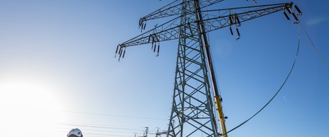 Der obere Teil eines Hochspannungsmasts hängt vor blauem Himmel an einem Kran, ein Arbeiter hält ein am Mast befestigtes Kabel Der obere Teil eines Hochspannungsmasts hängt vor blauem Himmel an einem Kran, ein Arbeiter hält ein am Mast befestigtes Kabel