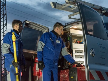 Zwei Techniker in blauen Overalls laden vor einer Abendstimmung Geräte aus einem Fahrzeug Zwei Techniker in blauen Overalls laden vor einer Abendstimmung Geräte aus einem Fahrzeug