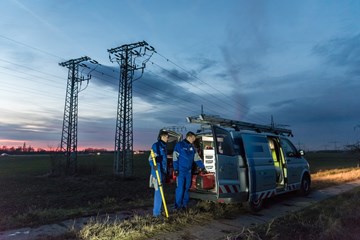 Zwei Techniker in blauen Overalls laden in der Dämmerung auf einem Feld mit Hochspannungsmasten Geräte aus einem Fahrzeug Zwei Techniker in blauen Overalls laden in der Dämmerung auf einem Feld mit Hochspannungsmasten Geräte aus einem Fahrzeug