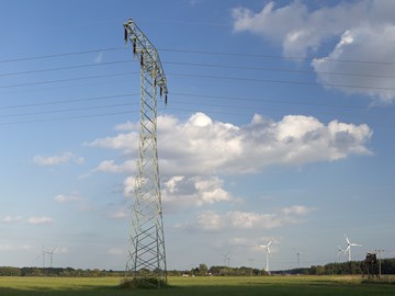 Hochspannungsmast auf einer grünen Wiese mit Windrädern unter blauem Himmel mit weißen Wolken Hochspannungsmast auf einer grünen Wiese mit Windrädern unter blauem Himmel mit weißen Wolken