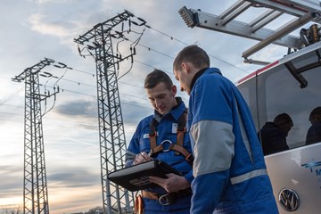 Zwei Monteure in Blaumännern blicken, am Heck eines Fahrzeugs vor zwei Hochspannungsmasten stehend, auf ein portables Eingabegerät Zwei Monteure in Blaumännern blicken, am Heck eines Fahrzeugs vor zwei Hochspannungsmasten stehend, auf ein portables Eingabegerät