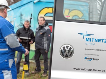 Zwei Männer in Parkern mit einem Arbeiter im Blaumann neben einer geöffneten Fahrzeugtür mit MITNETZ STROM-Logo Zwei Männer in Parkern mit einem Arbeiter im Blaumann neben einer geöffneten Fahrzeugtür mit MITNETZ STROM-Logo