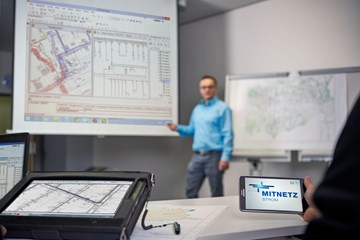 Laptop, portables Eingabegerät und Smartphone mit MITNETZ STROM-Logo - im Hintergrund hält ein Redner vor einem Plan seine Präsentation Laptop, portables Eingabegerät und Smartphone mit MITNETZ STROM-Logo - im Hintergrund hält ein Redner vor einem Plan seine Präsentation