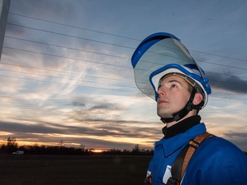Monteur mit Helm und geöffnetem Schutzvisier vor einem Hochspannungsmast in der Dämmerung Monteur mit Helm und geöffnetem Schutzvisier vor einem Hochspannungsmast in der Dämmerung