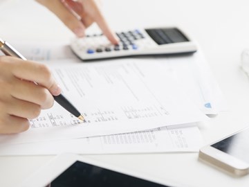 Hände mit Kugelschreiber und Taschenrechner über Unterlagen auf einem weißen Tisch Hände mit Kugelschreiber und Taschenrechner über Unterlagen auf einem weißen Tisch