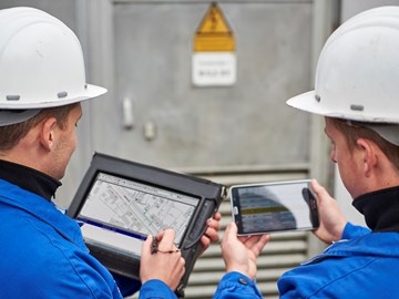 Zwei Techniker betrachten Pläne und Daten auf einem portablen Eingabegerät und einem Tablet nebeneinander Zwei Techniker betrachten Pläne und Daten auf einem portablen Eingabegerät und einem Tablet nebeneinander