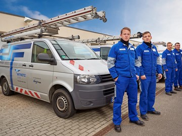 Vier Mitarbeiter in blauer Arbeitskleidung stehen vor einer Reihe aus geparkten Technikfahrzeugen im Freien Vier Mitarbeiter in blauer Arbeitskleidung stehen vor einer Reihe aus geparkten Technikfahrzeugen im Freien