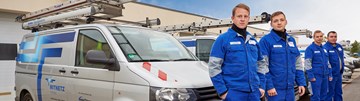 Vier Mitarbeiter in blauer Arbeitskleidung stehen vor einer Reihe aus geparkten Technikfahrzeugen im Freien Vier Mitarbeiter in blauer Arbeitskleidung stehen vor einer Reihe aus geparkten Technikfahrzeugen im Freien