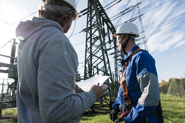 Zwei Männer mit weißen Helmen stehen unter Hochspannungsmasten, einer trägt eine Kletterausrüstung Zwei Männer mit weißen Helmen stehen unter Hochspannungsmasten, einer trägt eine Kletterausrüstung